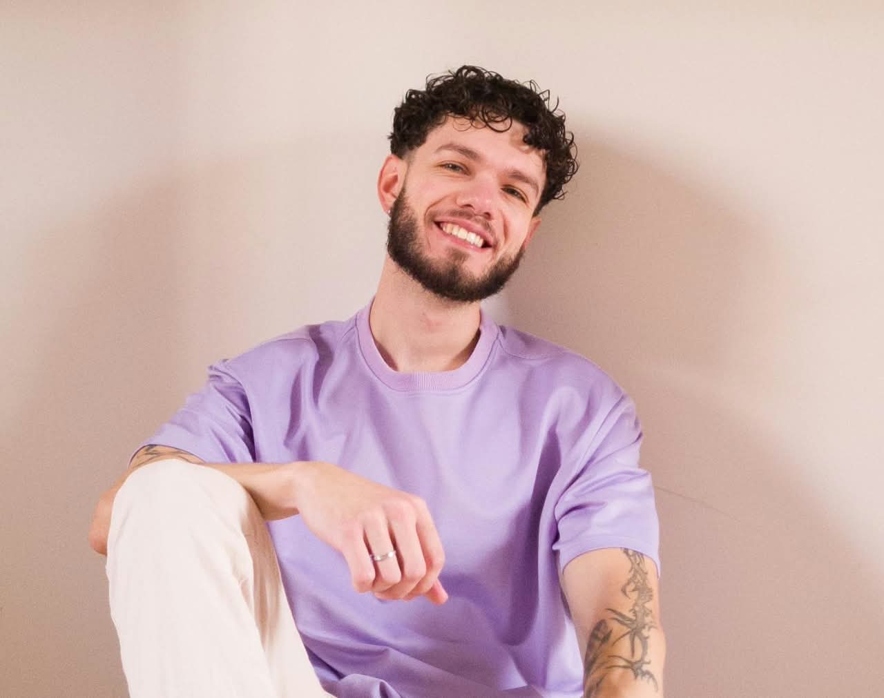 Retrato de Lucas Munhoz Gonçalves sorrindo, de camiseta lilás, diante de uma parede neutra