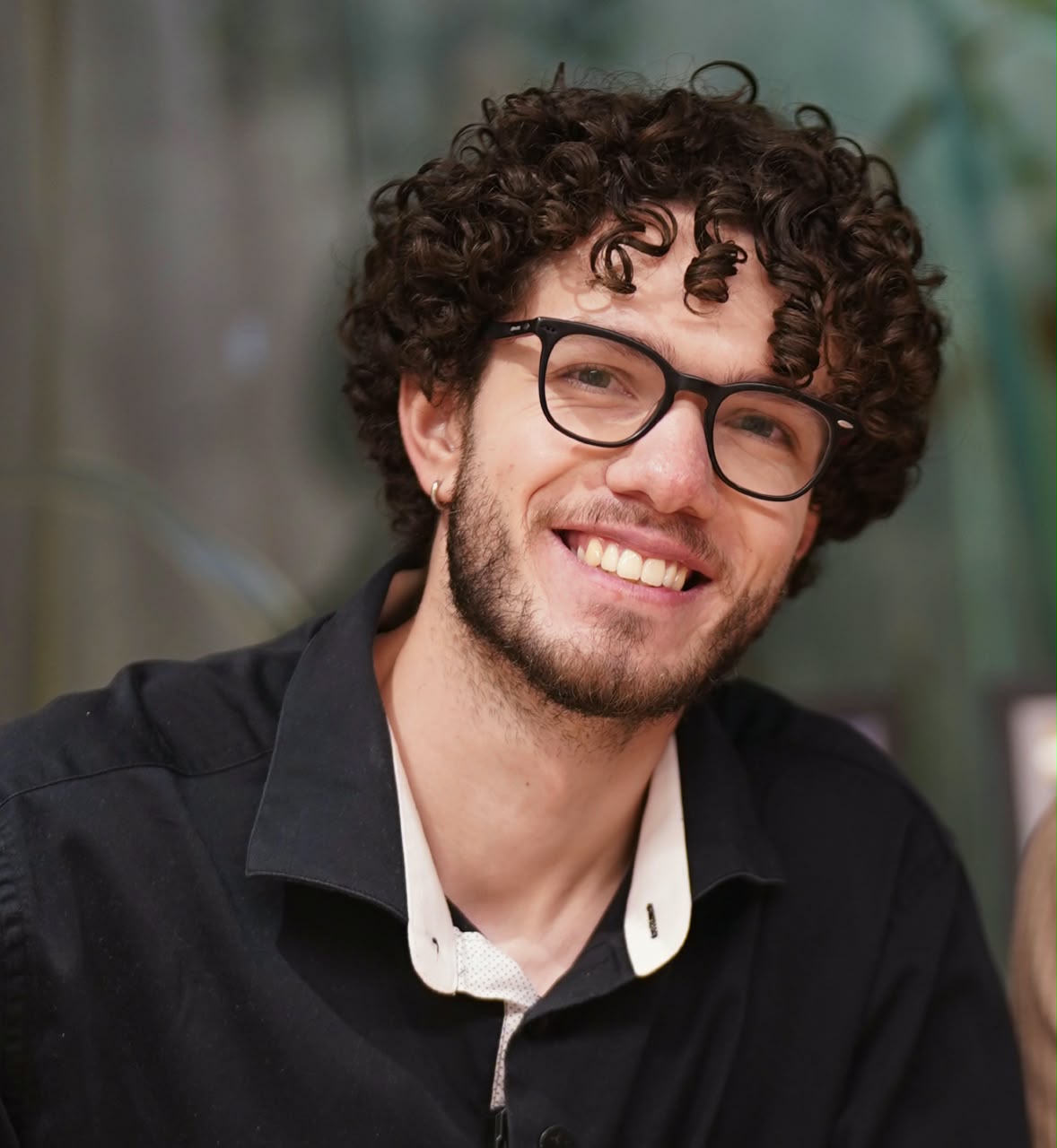 Retrato de Lucas Munhoz Gonçalves, psicólogo, sorrindo, de óculos e camisa escura